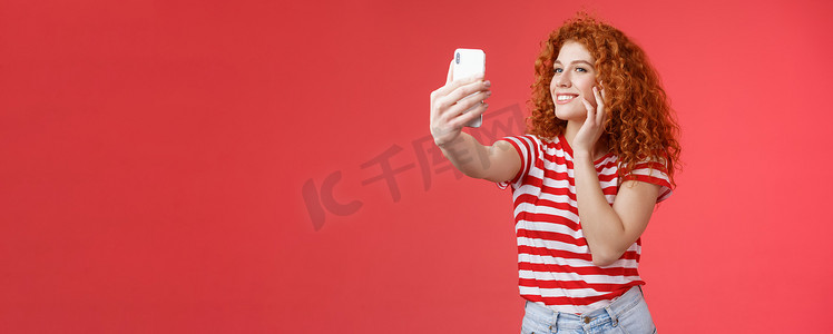流行开朗好看的时尚女博主红发卷发感觉很自我接受自拍举起手臂拿着智能手机摆出愚蠢可爱的手机摄像头红色背景