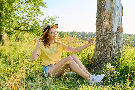 手机夏季夏季摄影照片_戴草帽的成年女性放松，坐在草地上，看着智能手机网络摄像头，打视频电话