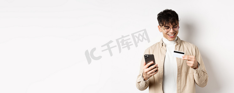购物app摄影照片_戴眼镜的快乐年轻人在网上购物，看着塑料信用卡，拿着智能手机，站在白色背景上