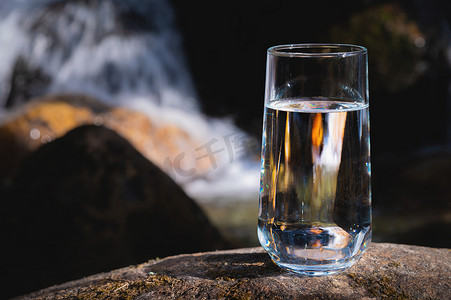 在大自然森林中的户外石头上，玻璃杯中饮用透明水，瀑布在模糊的背景中肆虐