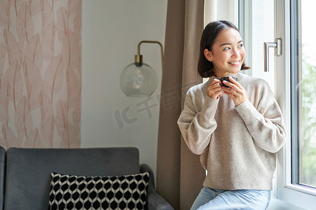 路人摄影照片_美丽的亚洲女人坐在家里喝杯咖啡，一边享用浓缩咖啡，一边看着窗外的路人，开心地笑着