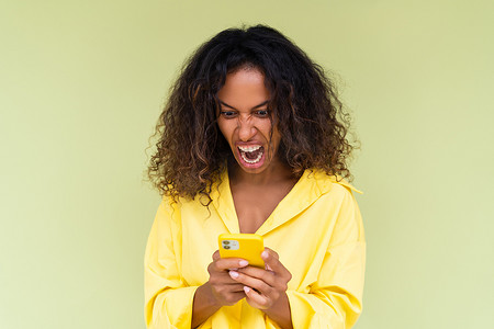 绿色背景中身穿休闲衬衫的美丽非洲裔美国女性愤怒地拿着手机看着屏幕