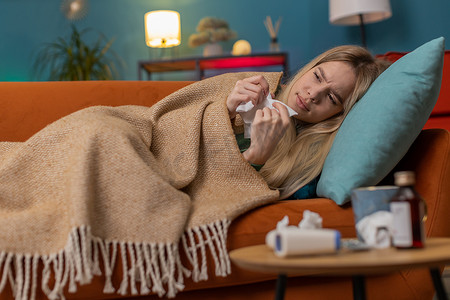 生病的年轻女子患感冒、过敏，躺在家里的沙发上打喷嚏，把鼻涕擦进餐巾纸