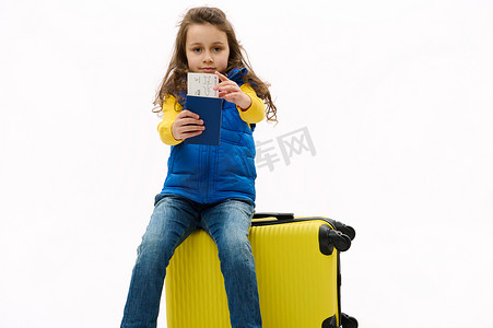 可爱的小女孩拿着机票和登机牌对着镜头，坐在黄色旅行包上，去周末度假