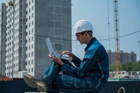 戴安全帽的白人男性建筑工人坐在楼板上，在建筑工地使用笔记本电脑。