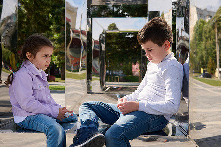 可爱的孩子们，兄弟姐妹坐在城市公园的镜子长椅上，在家庭出游时看起来很伤心。