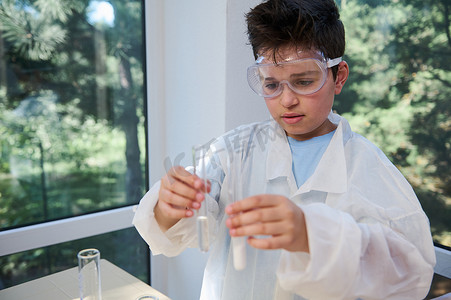 穿着白色实验室外套的青少年男学生拿着带化学实验室的试管。