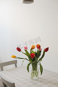 切花摄影照片_新鲜的春天五颜六色的郁金香花束站在白色桌子上的花瓶中，在浅色经典设计的客厅背景中。