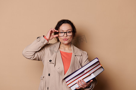 自信的女教育家，老师拿着一叠书，透过眼镜看着相机，米色背景