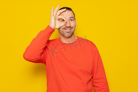 黄色手指手势摄影照片_40 多岁的留着胡子的西班牙裔男子穿着米色高领毛衣，用手指做一个 ok 的手势，孤立在黄色工作室背景中。