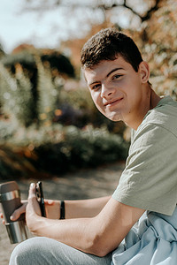 一个少年坐在秋天公园的长椅上，喝着保温杯里的咖啡，看着手机。