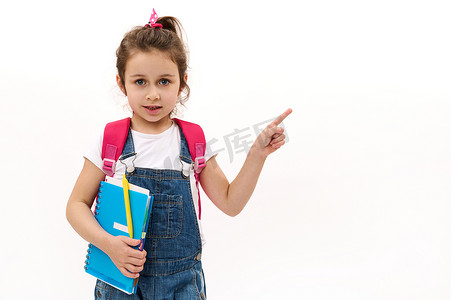新的学期摄影照片_小学生，可爱的女孩，带着抄写本和背包，用食指指着白色的复印空间。