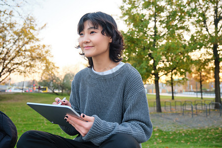 年轻微笑的韩国女孩、平面设计师、艺术家用钢笔工具在数字平板电脑上画画的肖像，坐在公园里呼吸新鲜空气，捕捉，做笔记