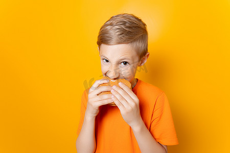 一个穿着橙色 T 恤的狡猾男孩咬着汉堡包，看着黄色背景中的镜头