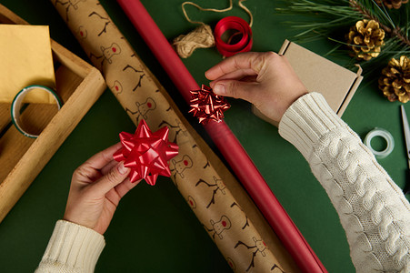 顶视图女人的手在绿色表面的包装纸上系上蝴蝶结，上面有包装圣诞礼物的材料