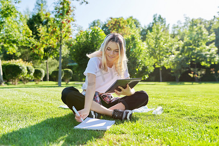 戴耳机的青少年学生坐在草坪上，在笔记本上写字