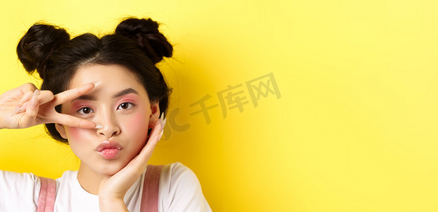 特写：可爱的亚洲女性，妆容艳丽，留着长长的指甲，在镜头前展示 V 形标志，傻傻地噘着嘴，站在黄色背景中的卡哇伊