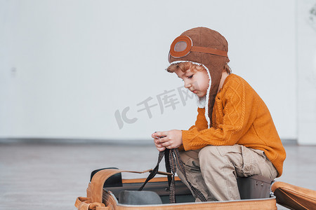 白天，穿着复古飞行员服装的小男孩玩得很开心，坐在室内的手提箱里