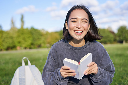 读书器摄影照片_美丽的亚洲女孩坐在公园的草地上，边读书边微笑。