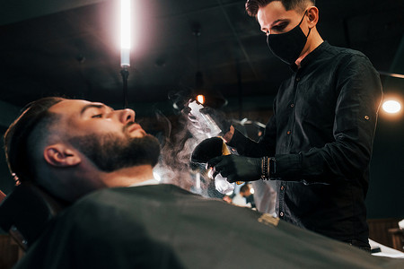 剃胡子的男人摄影照片_发型时髦的年轻人坐着，在理发店被戴黑色防护面具的男人剃胡子