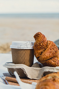 咖啡原料摄影照片_美味的新鲜羊角面包和沙滩海岸背景纸杯中的咖啡。