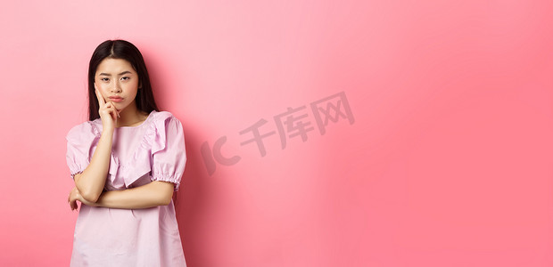 无聊的亚洲少女对着镜头漠不关心，手托着脸，摆出怀疑的姿势，站在粉红色背景下不情愿