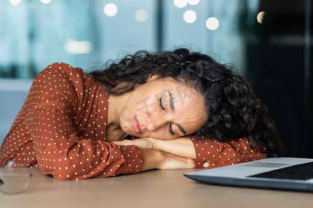 睡在办公桌上的美丽疲惫的西班牙裔女性，闭着眼睛在笔记本电脑附近的办公室里打盹的特写女商人