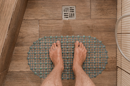 男人的脚站在浴室或淋浴间地漏旁边的塑料防滑垫上