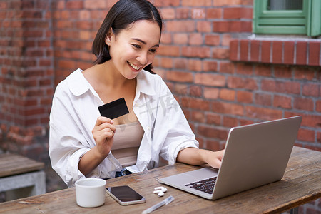 微笑的亚洲女性坐在咖啡馆里，拿着笔记本电脑，用信用卡支付网上购物费用，发送她的银行账户详细信息