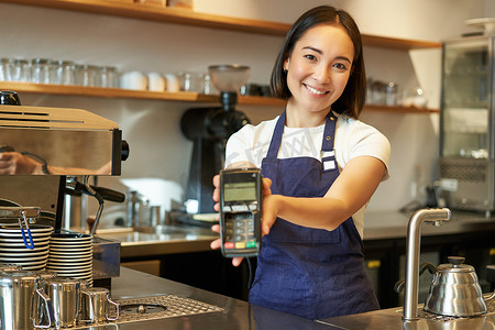 微笑的亚洲咖啡师，咖啡店员工给你信用卡机，用 POS 终端处理付款，在咖啡馆工作