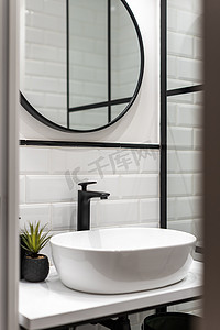 白色砖墙的浴室，黑色水龙头，白色桌子上的椭圆形洗脸盆，黑色镜框墙上的圆形镜子。