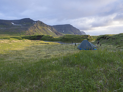 小蓝色帐篷独自站在 Hornstrandir 冰岛长满苔藓的溪岸上的绿草上，积雪覆盖的山丘和悬崖，多云的天空背景，黄金时段的灯光，复制空间