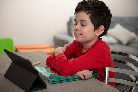 可爱的十几岁男孩，聪明的小学生坐在桌边，在数字平板电脑上享受在线课程