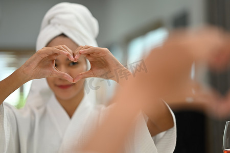 自信的千禧一代穿着浴袍的女人用手做心形符号，对着镜子里的倒影微笑