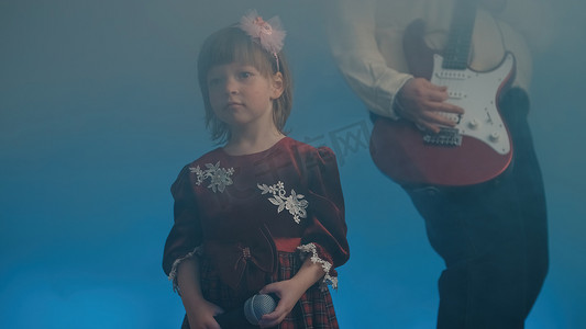 家庭舞台摄影照片_穿着复古裙子的小女孩在舞台上唱歌，她的父亲弹着电吉他