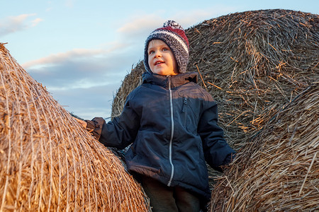 傍晚的阳光下，一个戴着针织帽的微笑可爱男孩站在金色的稻草堆上。