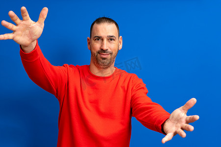 一个留着胡子的拉丁裔男子的画像，身穿红色套头衫，张开双臂欢迎，与蓝色工作室背景隔离。