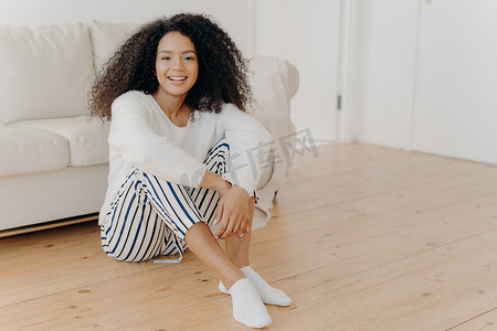 室内拍摄的宁静快乐的非洲裔美国女性坐在客厅的地板上，背景是白色沙发，穿着时髦的衣服和袜子，表达积极的情绪，独自在家休息