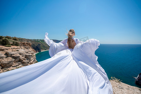 金发碧眼的长发在阳光明媚的海边穿着白色飘逸的连衣裙，后视，丝绸面料在风中飘扬。
