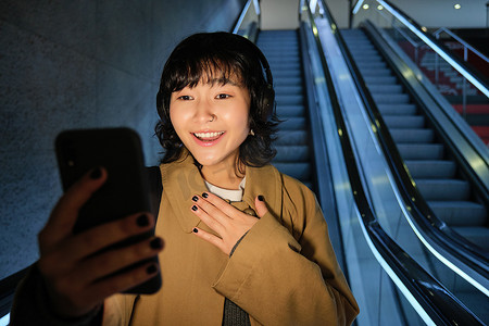 亚洲女孩的肖像使用智能手机，戴着耳机听音乐，晚上从市中心的自动扶梯上下来，看着手机很惊讶，惊讶地喘着粗气