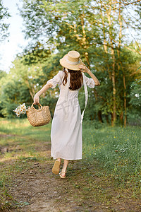 一位身穿夏装长裙的女士在林间小径上行走，手上戴着一顶柳条帽