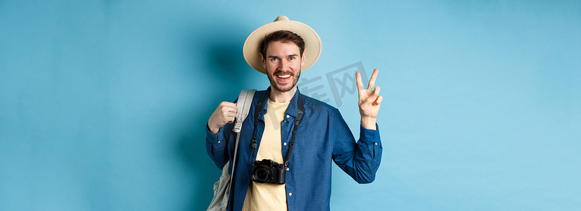 快乐帅哥暑假拍照，展示和平标志，微笑，戴着草帽，拿着旅游背包，蓝色背景