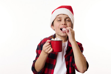 戴圣诞帽的男孩摄影照片_快乐的男孩，戴着圣诞帽的少年，享用热巧克力饮料，吃着白色背景中美味的甜棉花糖