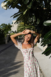 在热带树叶的背景下，身着浪漫连衣裙、带花卉图案和珍珠项链手链的年轻美女