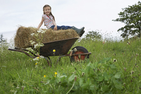 坐在推车的女孩摄影照片_女孩（5-6 岁）坐在手推车的干草上，在田野肖像中