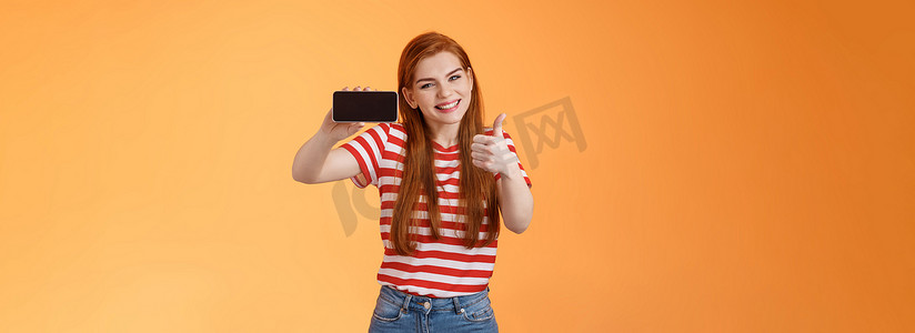 兴奋开朗的红发美女很高兴，竖起大拇指，介绍很棒的手机游戏，水平握住智能手机，微笑表示认可，推荐好的网上商店，橙色墙