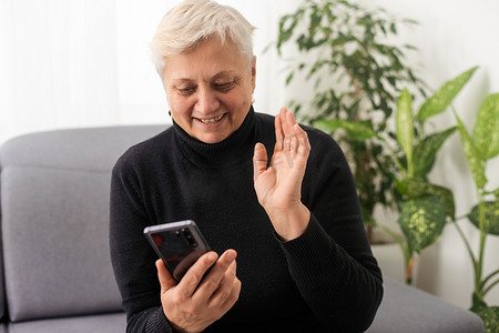 移动端摄影照片_轻松成熟的 60 多岁老妇人、年长的中年女性顾客手持智能手机使用移动应用程序、短信、搜索电子商务提供手机技术设备坐在家里的沙发上