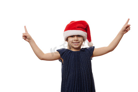 戴着圣诞帽的快乐女婴和时髦优雅的蓝色海军裙，可爱地微笑着，指着白色的复制空间