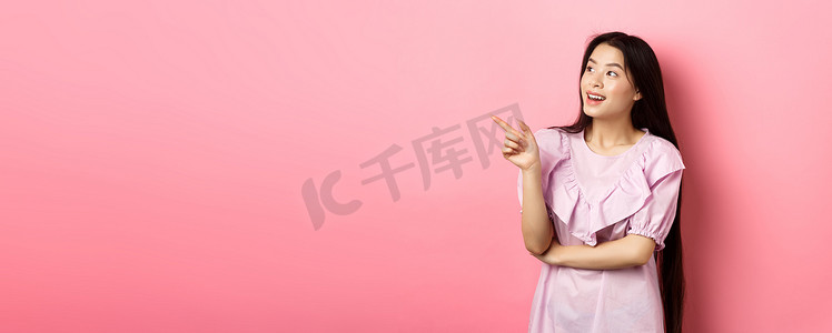 穿着裙子的兴奋微笑的亚洲女性，指着并向左看标志，查看促销，站在粉红色背景中