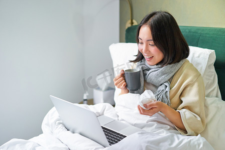 亚洲生病女孩的画像，因感冒躺在床上，患流感，与笔记本电脑交谈，视频聊天，在病假期间在家远程工作